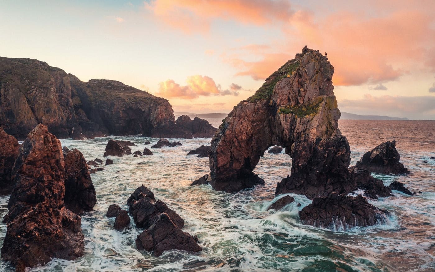 A large, jagged sea arch rises from the rocky coastline, surrounded by crashing waves, with a pastel-colored sunset sky in the background.