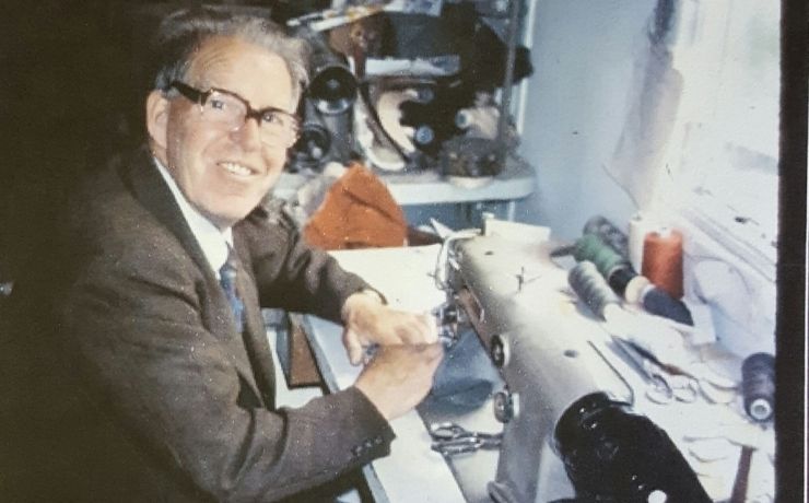 An older man wearing glasses and a suit smiles while sewing fabric on an industrial sewing machine in a workshop filled with thread spools and sewing tools.