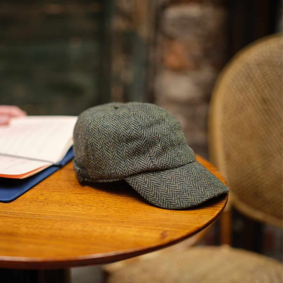 A gray herringbone-patterned cap rests on a round wooden table next to an open book, with two woven chairs and a blurred brick wall in the background.