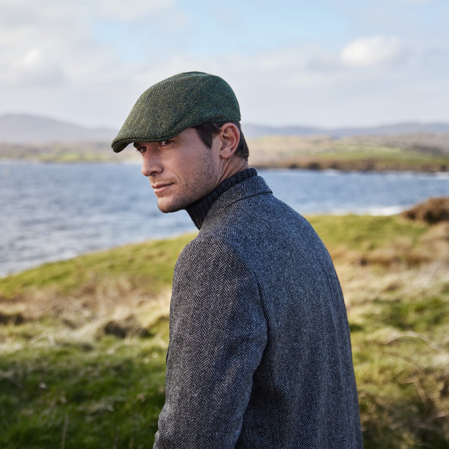 A man wearing a gray herringbone coat and a green flat cap stands outdoors near a body of water, looking over his shoulder. The landscape is grassy with rolling hills and a cloudy sky in the background.