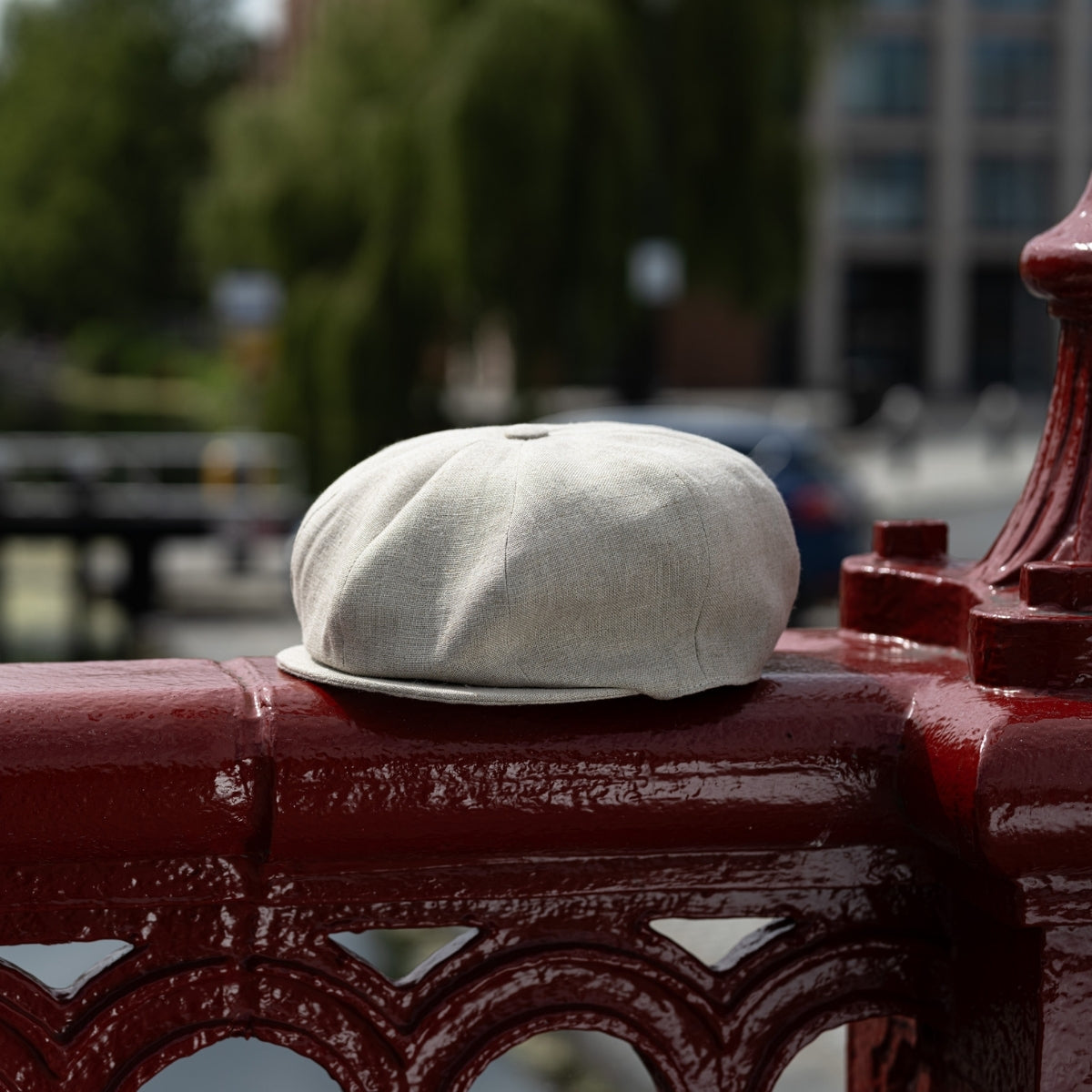 A light gray flat cap rests on a glossy red railing outdoors, with blurred trees, buildings, and a street in the background.