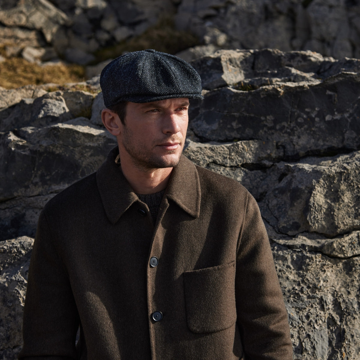 A man wearing a dark flat cap and brown coat stands in front of large, rugged gray rocks, looking slightly to the side under natural sunlight.