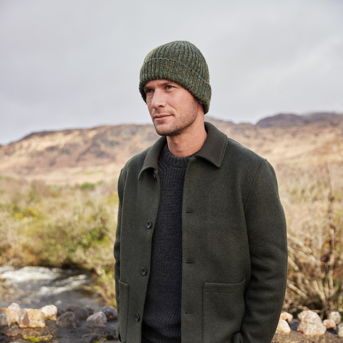 A man wearing a green knit beanie and dark green coat stands outdoors with his hands in his pockets. Behind him are hills, a stream, and cloudy sky, suggesting a rural, natural landscape.