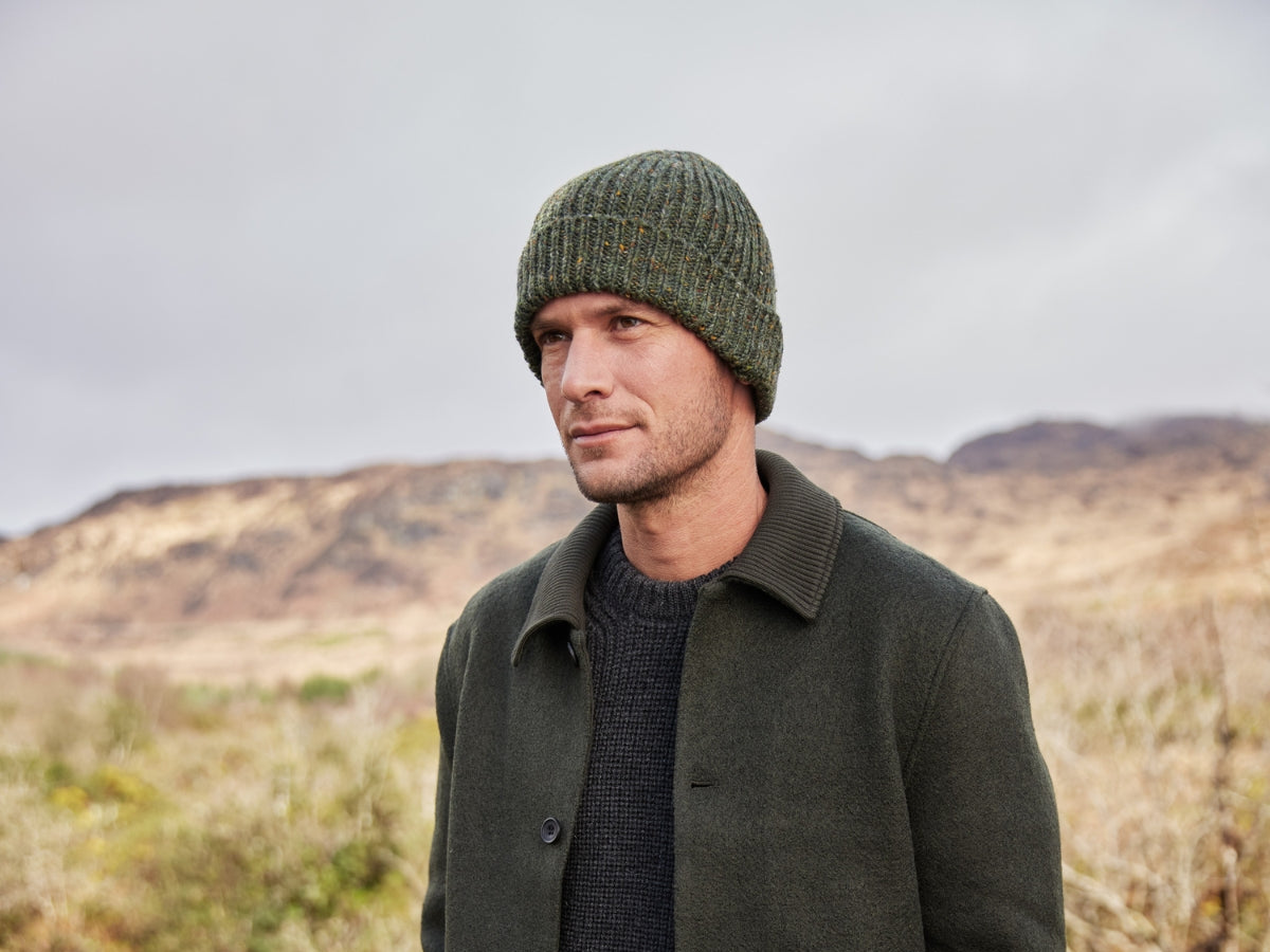 A man wearing a green knit beanie and dark green coat stands outdoors with his hands in his pockets. Behind him are hills, a stream, and cloudy sky, suggesting a rural, natural landscape.
