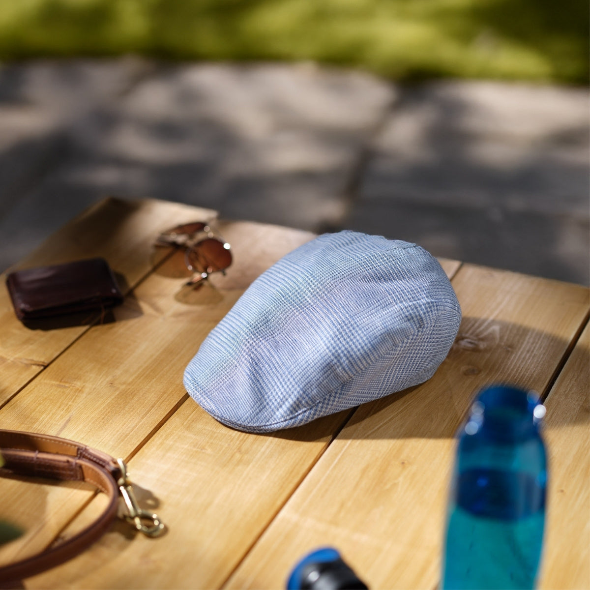 A light blue plaid flat cap rests on a wooden table outdoors, surrounded by sunglasses, a brown wallet, a dog leash, and a blue bottle. Sunlight and shadows create a relaxed, summery atmosphere.
