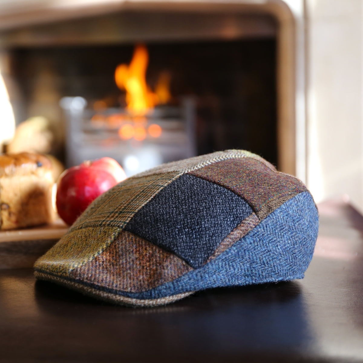 A patchwork flat cap made of various wool fabrics rests on a table, with a fireplace burning warmly in the background and an apple and bread nearby.