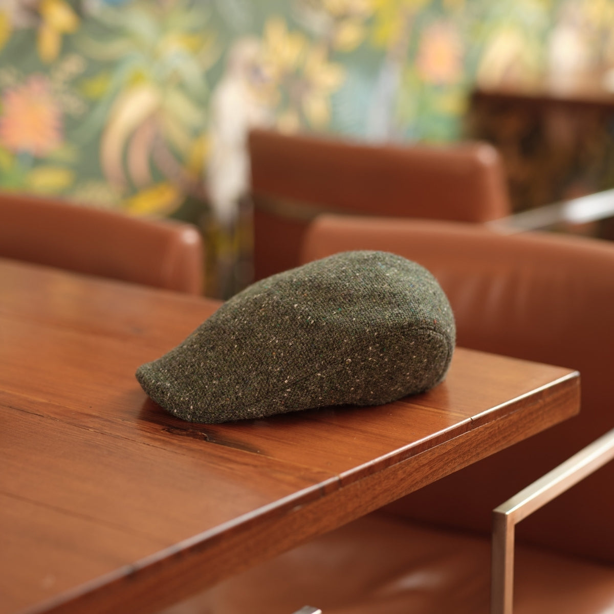 A green, textured fabric object shaped like a pear sits on a wooden table in a room with brown chairs and floral wallpaper in the background.