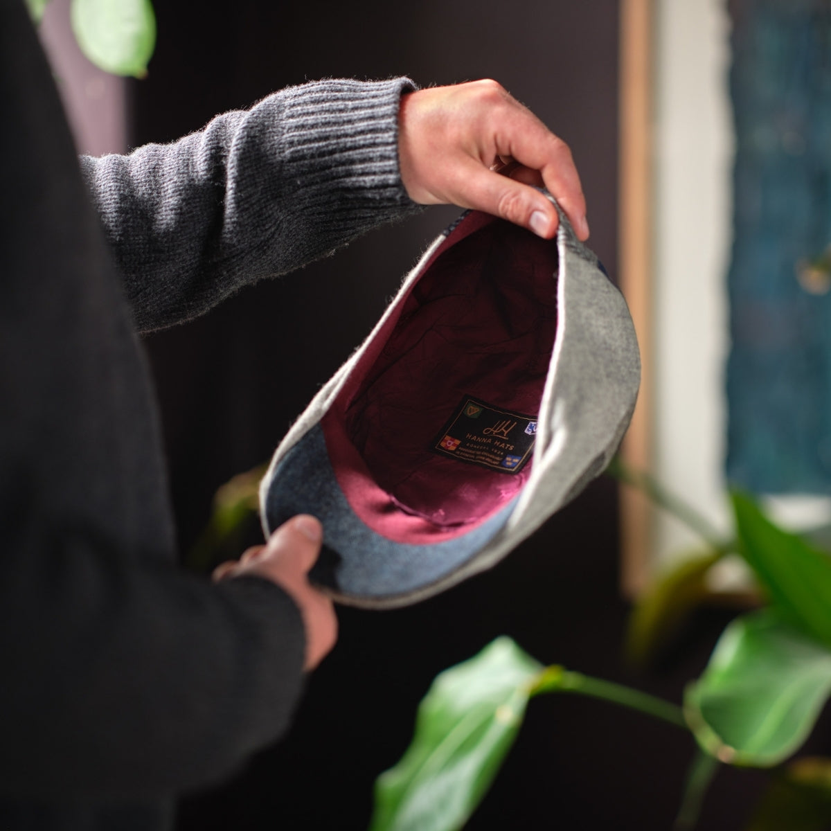 A person in a dark sweater holds a gray flat cap, showing its burgundy lining and an interior label. Green plants and a blurred background are visible.