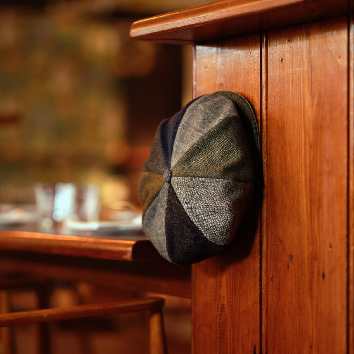 A patchwork flat cap in muted earth tones hangs on the corner of a polished wooden counter in a warmly lit, rustic restaurant.