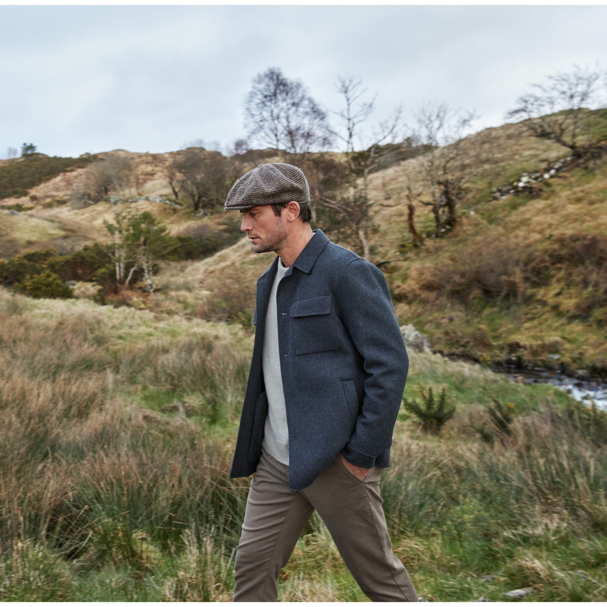 A man wearing a flat cap, grey jacket, and beige pants walks through a grassy, hilly landscape with sparse trees and a small stream in the background.