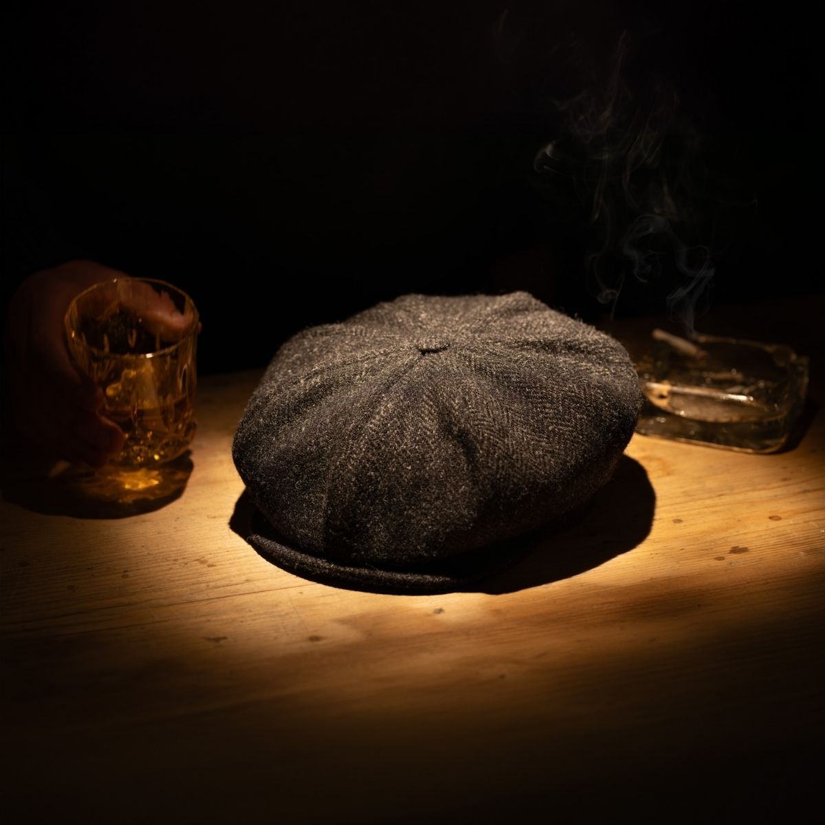 A flat cap is illuminated on a wooden table, beside a glass of whiskey held by a hand and a smoking cigarette in an ashtray, all surrounded by shadows.