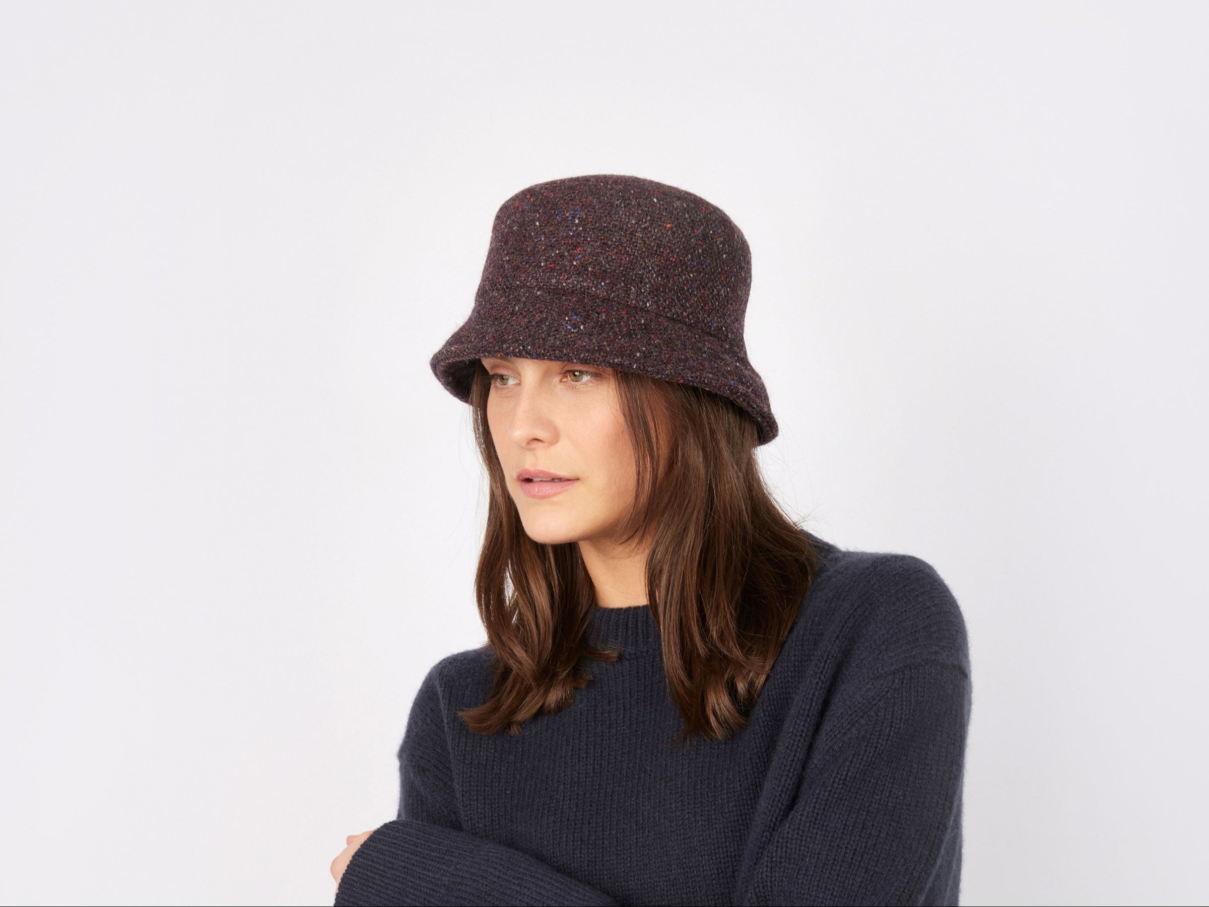 A woman with long brown hair wears the Eleanor Hat Tweed by Hanna Hats of Donegal Ltd and a navy sweater, standing against a plain white background.