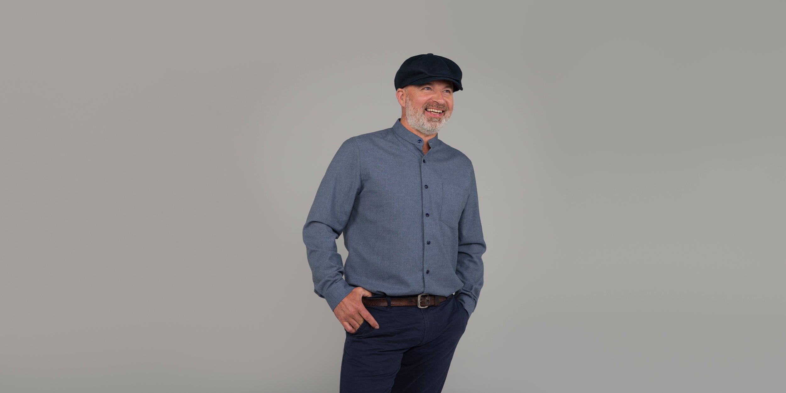 A smiling man with a gray beard, wearing a blue button-up shirt, dark pants, and a dark flat cap stands against a plain light gray background with one hand in his pocket.