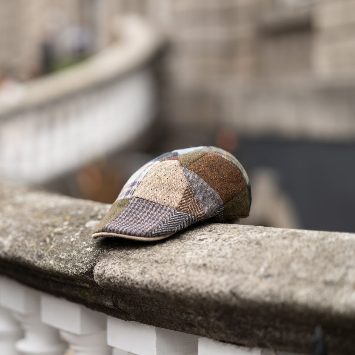 A patchwork flat cap made of various patterned fabrics rests on a curved stone railing, with a blurred background of steps and a stone building.