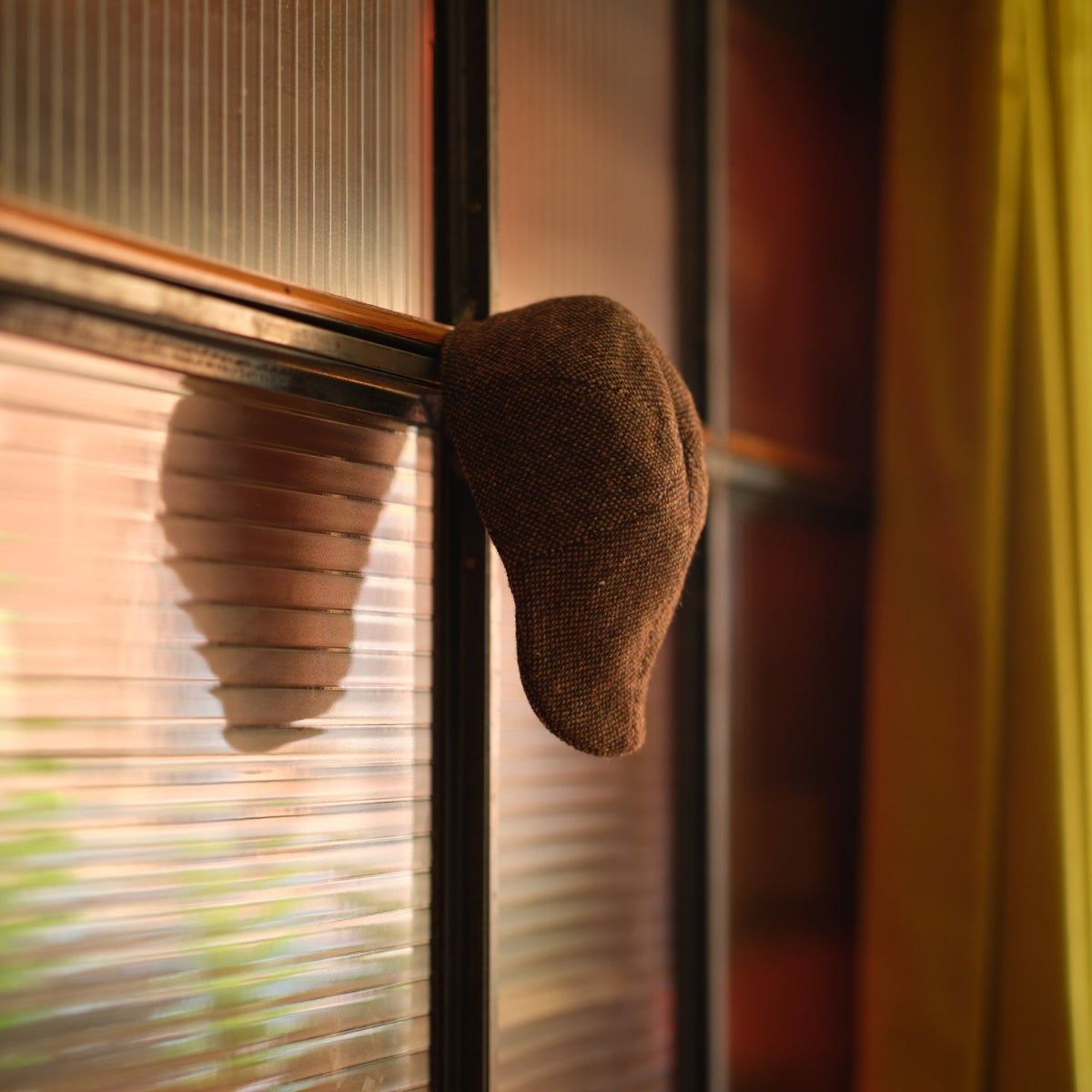 A brown flat cap hangs on the edge of a ribbed glass window, casting a shadow on the glass. Yellow curtains are visible to the right side of the image.