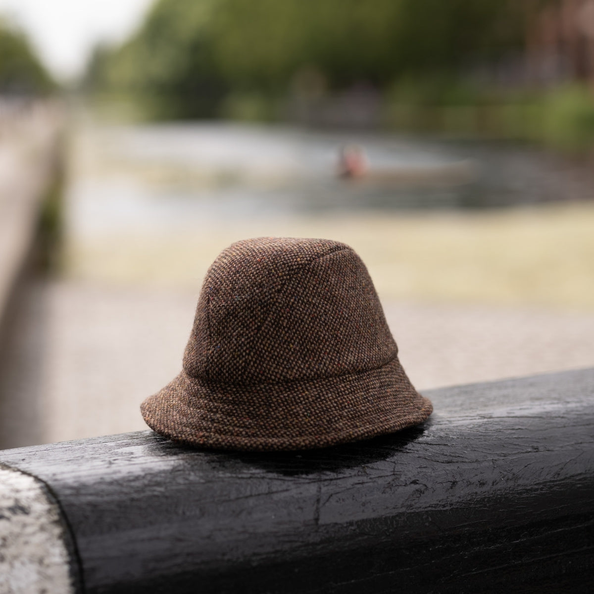 A brown tweed hat rests on a black railing beside a canal, with water and blurred greenery visible in the background.