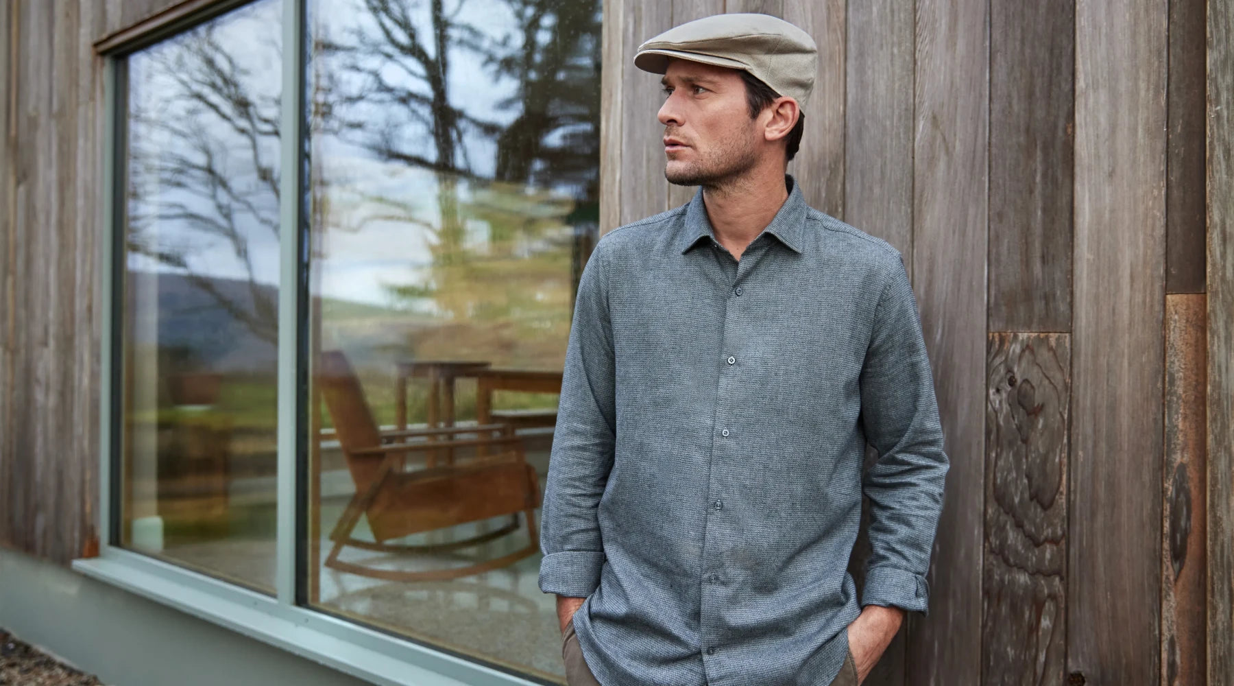 A man wearing a gray button-up shirt and beige flat cap stands with hands in his pockets in front of a wooden building with large windows reflecting trees and a rocking chair inside.
