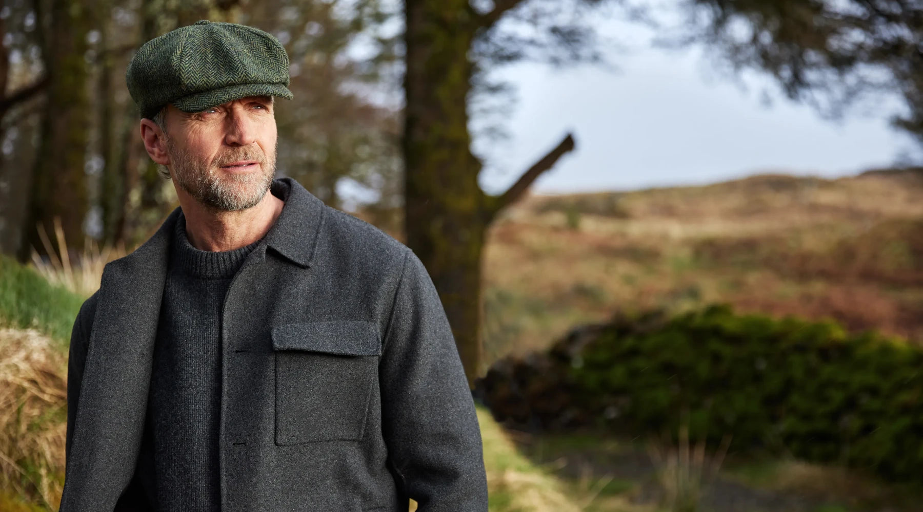 A bearded man wearing a flat cap and a dark coat stands outdoors in a rural, grassy landscape with trees and hills in the background, looking thoughtfully into the distance.