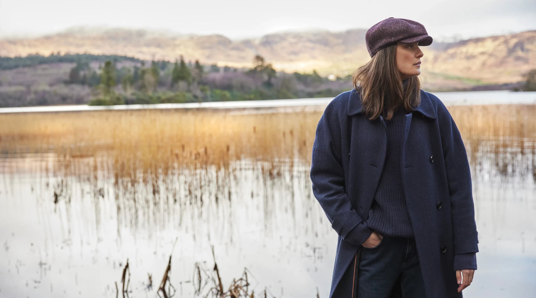 A woman wearing a dark blue coat, sweater, and a flat cap stands by a calm lake with reeds, with hills and trees in the background under a cloudy sky.