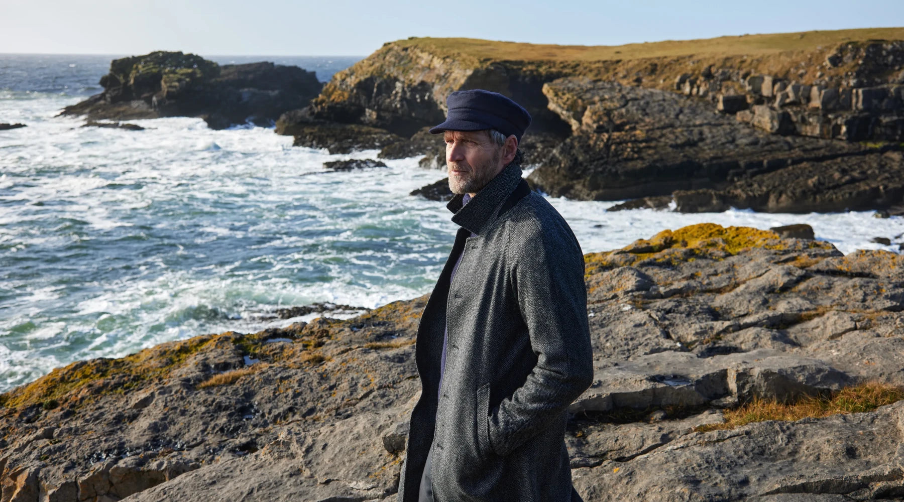 A man wearing a dark coat and navy cap stands on a rocky coastline, looking out toward the ocean. Waves crash against the rocks under a clear sky, and cliffs stretch along the shore in the background.