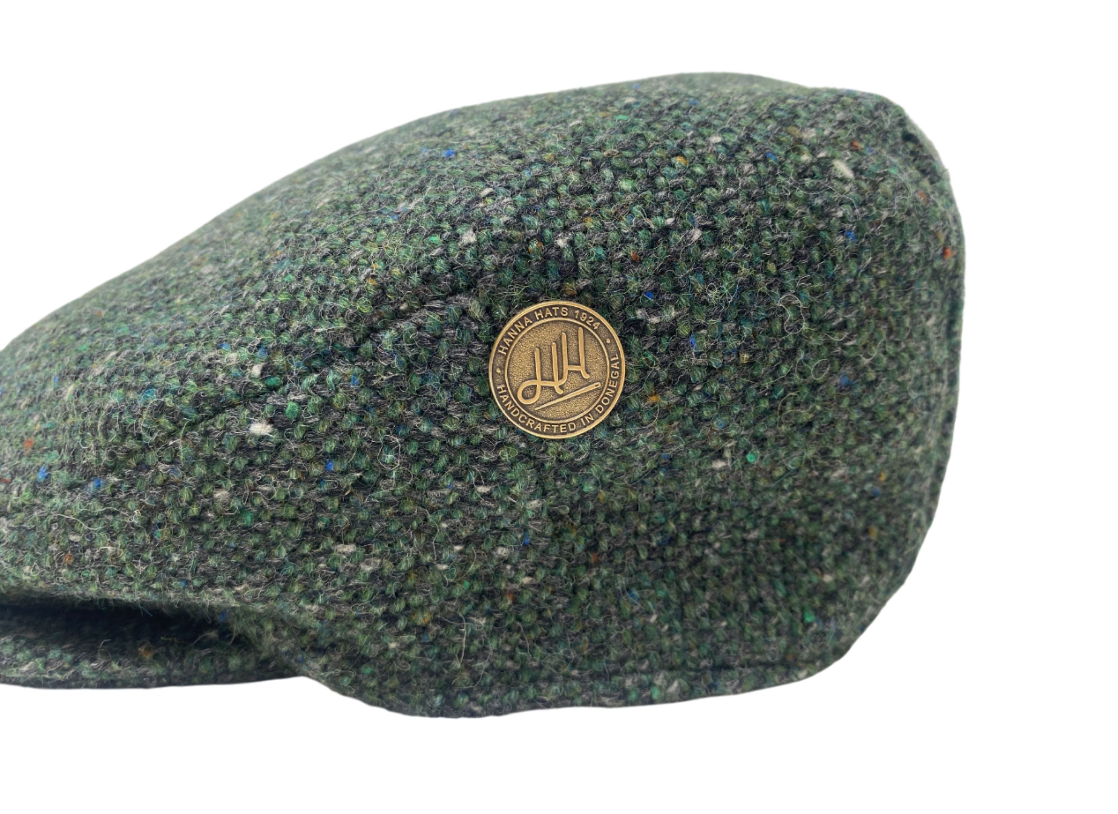 The Hanna Hats Pin by Hanna Hats of Donegal Ltd features a round logo with intricate detailing, shown on the side of a green tweed flat cap, displayed on a white background for a classic heritage look.