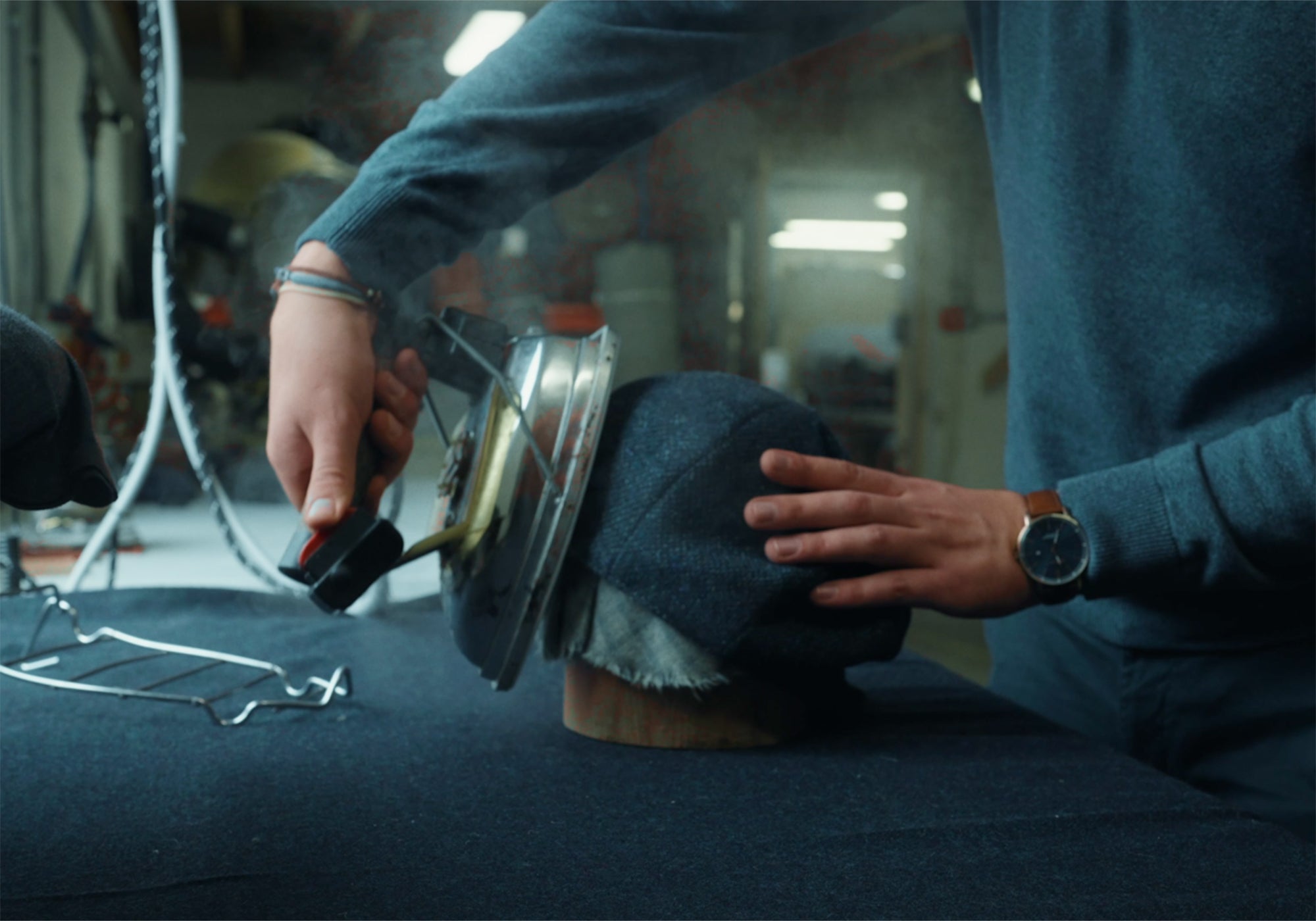 A person steams a dark fabric hat with a large iron, shaping it on a wooden mold. The person wears a dark blue sweater and a watch, working on a dark table in a workshop setting.