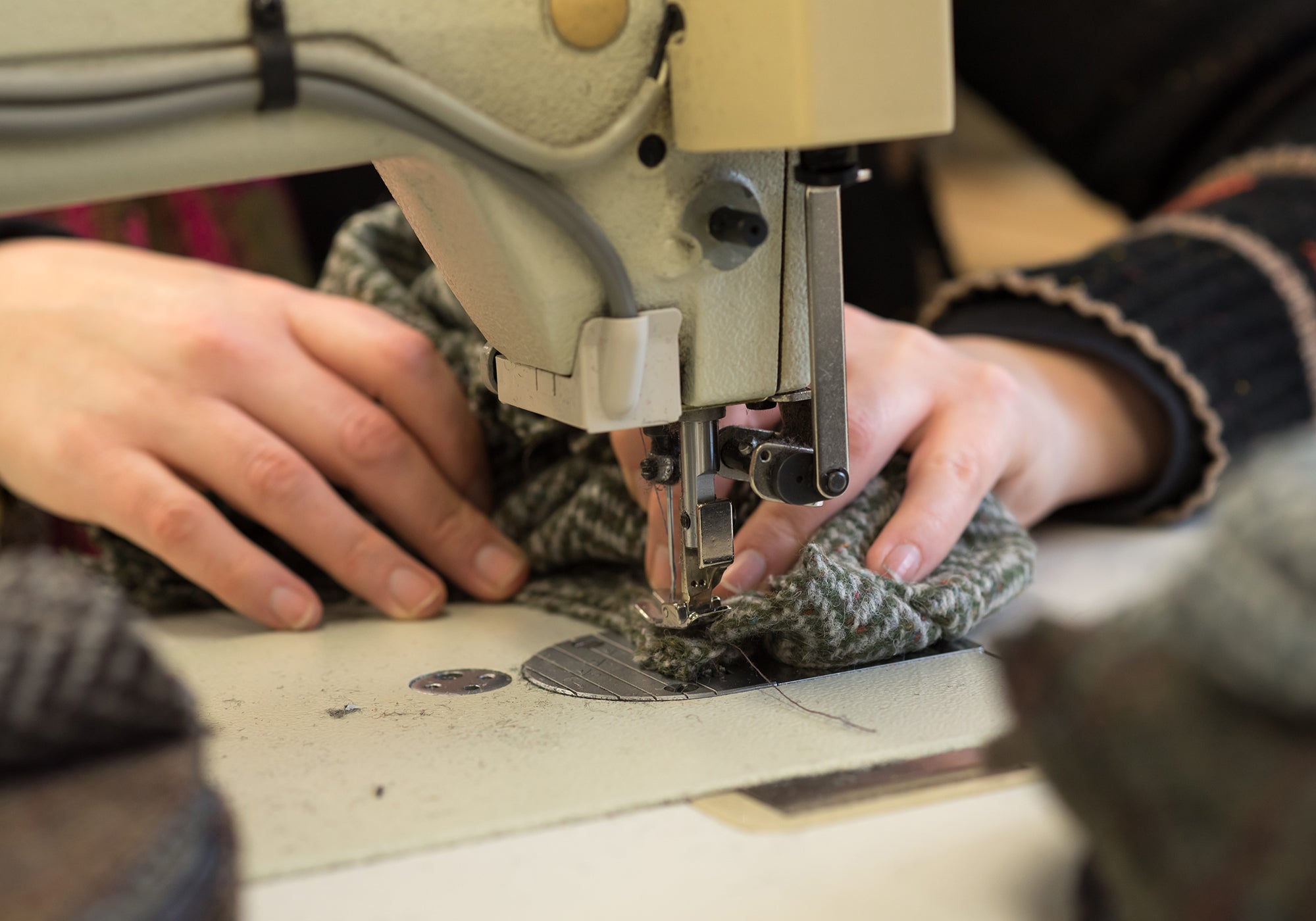 Close-up of hands guiding patterned fabric through a sewing machine, stitching the material together. The focus is on the needle and fabric, with other textile pieces blurred in the foreground.
