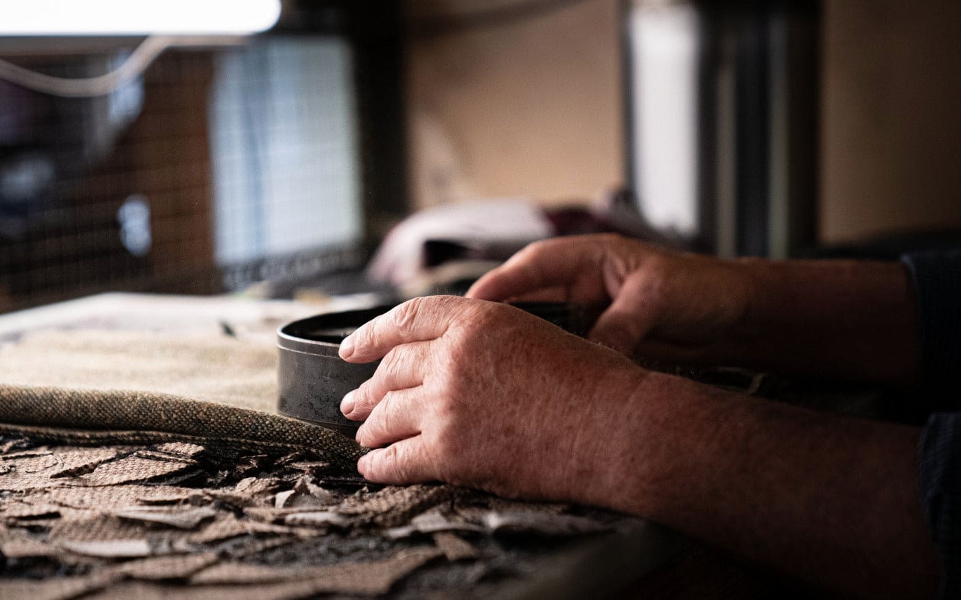 A close-up of a person's hands working with a round black container on a workbench covered with fabric and leather scraps, under bright lighting in a workshop setting.
