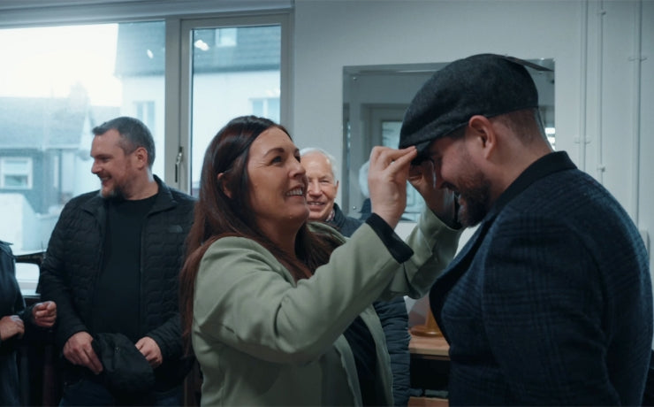 A woman smiles as she adjusts a flat cap on a man's head in a room with other people. Everyone appears cheerful and relaxed, standing near large windows with daylight outside.