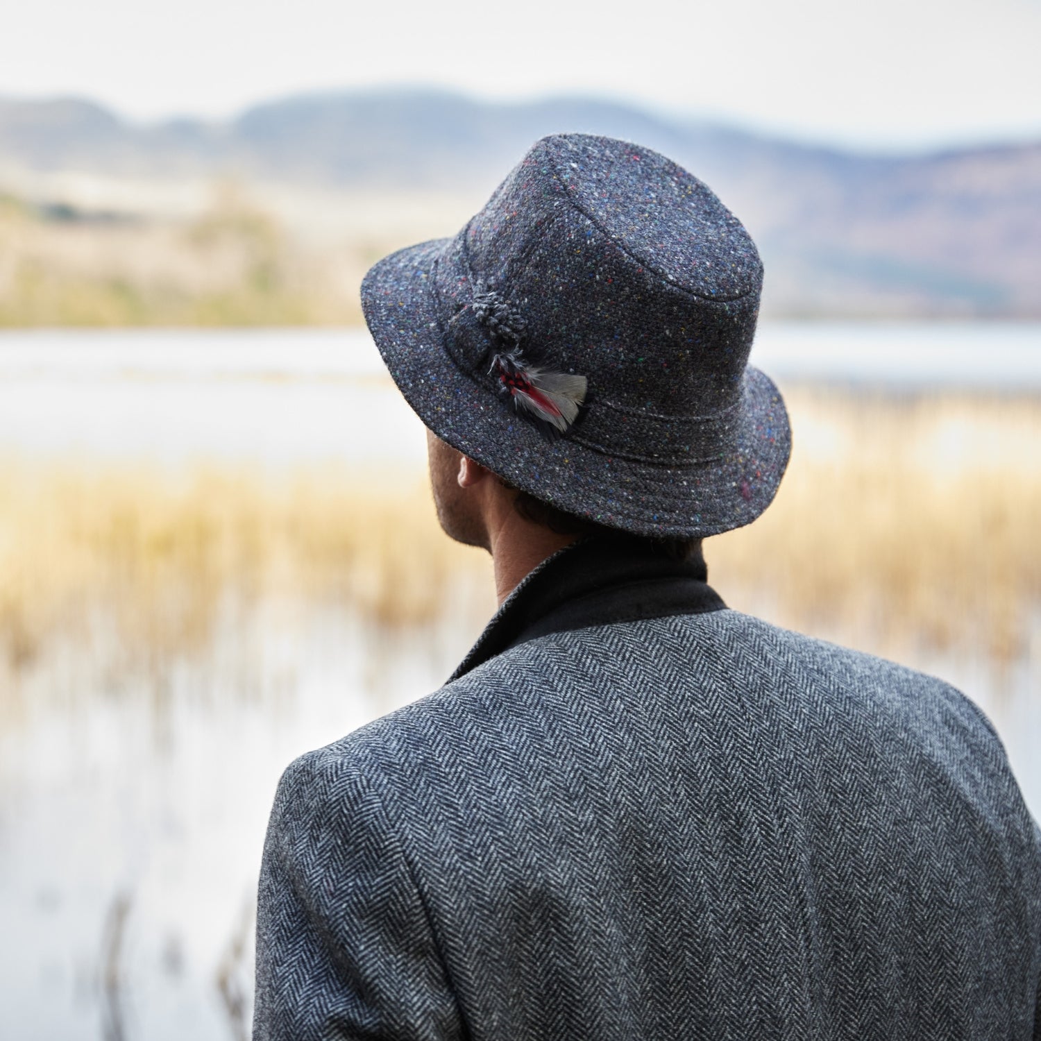 A person wearing a gray herringbone coat and a speckled gray hat with a feather looks out over a calm, reed-filled lake with hills in the background.