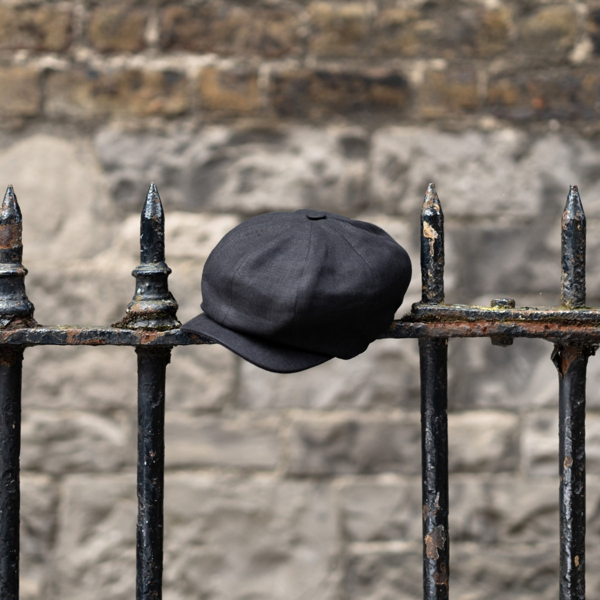 A flat cap rests on a black iron fence with pointed tips, set against a weathered stone wall in the background.