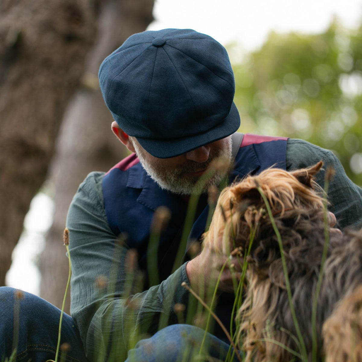 A man wearing a blue cap and vest sits outdoors, gently holding and looking at a small, long-haired dog among tall grass and trees.