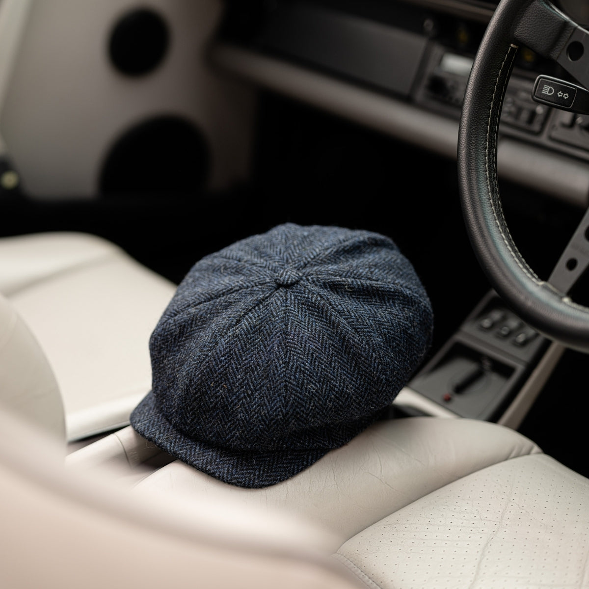 A dark blue herringbone flat cap rests on the light-colored leather seat of a car, with part of the steering wheel and dashboard visible.