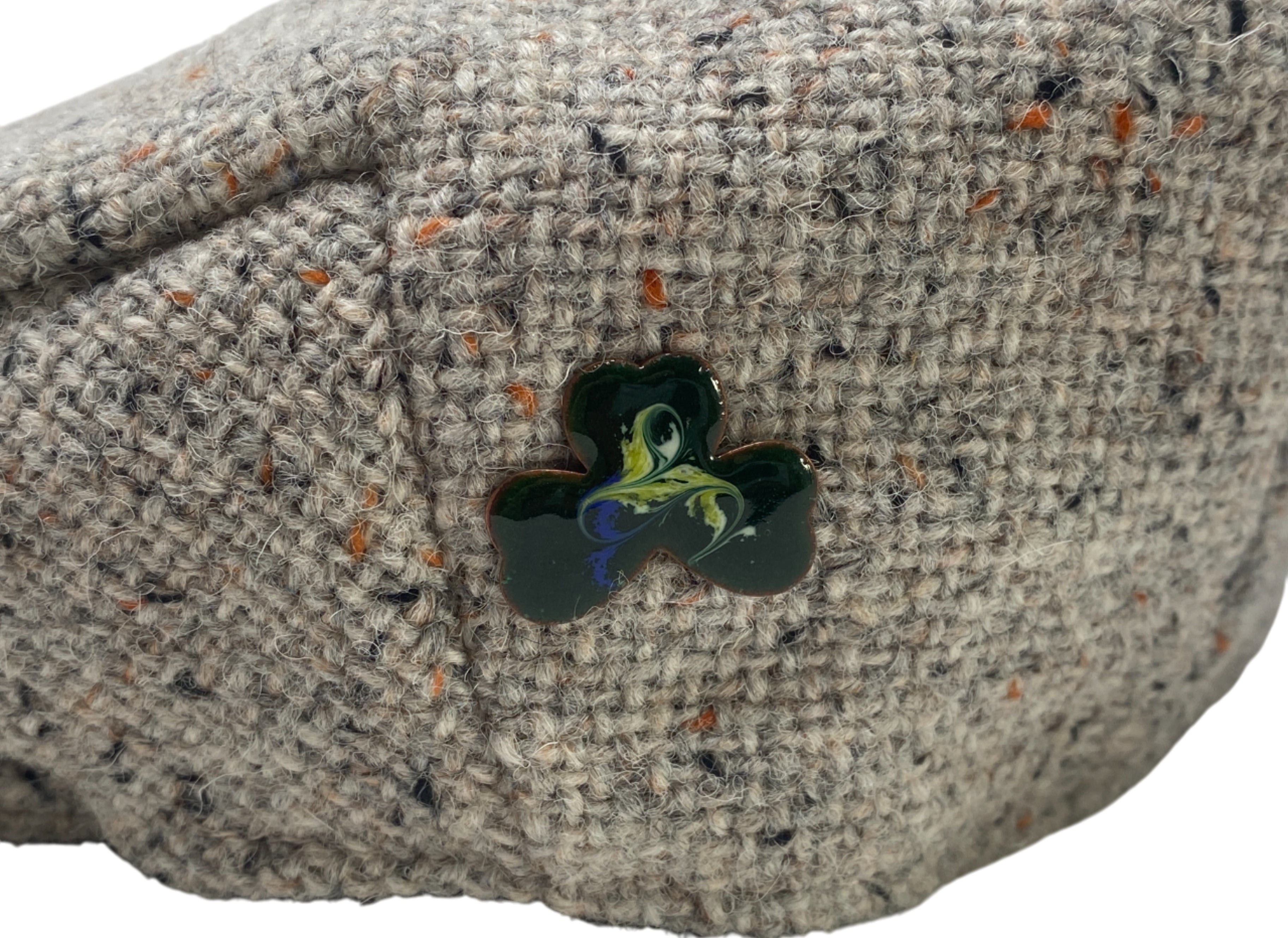 A close-up of a textured beige tweed cap by Hanna Hats of Donegal Ltd, featuring the Meab Enamels Lucky Shamrock Pendant—crafted from recycled copper with orange and black specks using open fire enamelling.