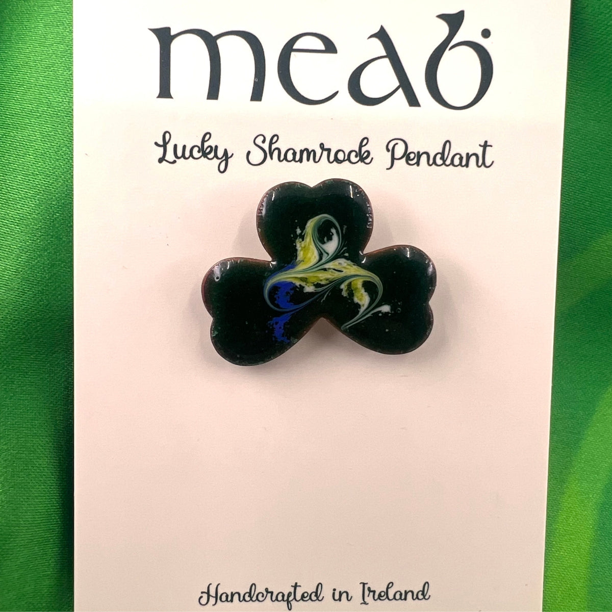 A black shamrock-shaped pendant with a yellow and green swirl design, displayed on a card labeled "meab Lucky Shamrock Pendant" and "Handcrafted in Ireland," against a green background.