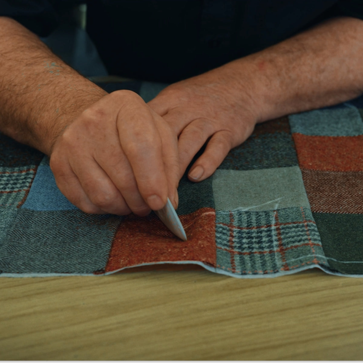 A person marks a patchwork fabric with tailor’s chalk on a wooden table, preparing it for sewing.