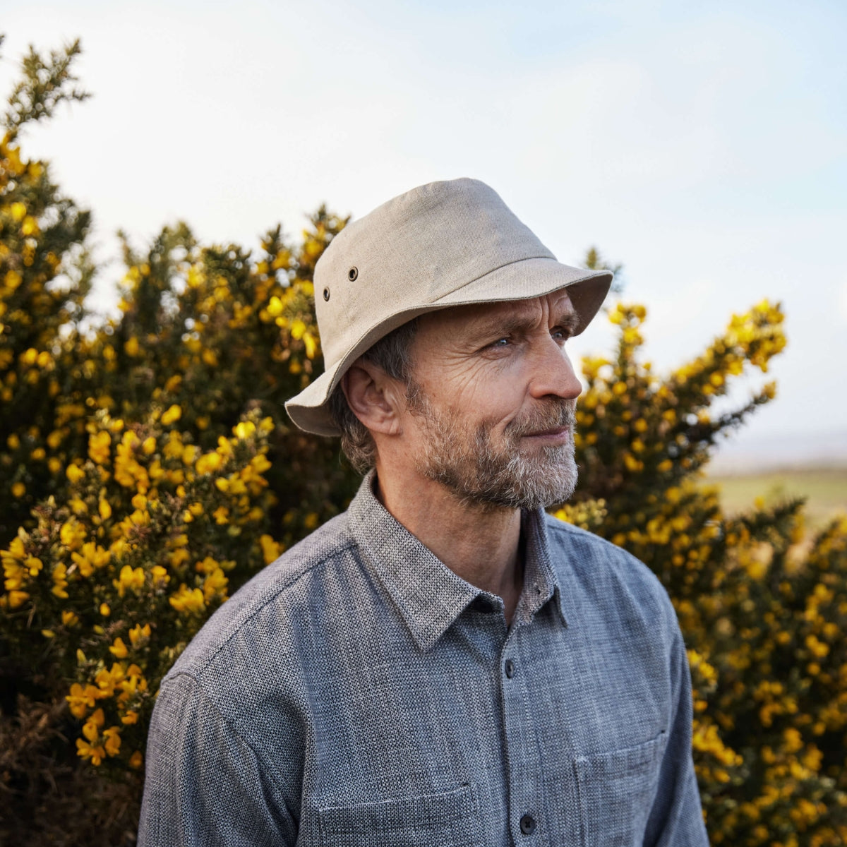 A middle-aged man with a beard wears a light-colored bucket hat and a gray button-up shirt, standing outdoors in front of green bushes with small yellow flowers.