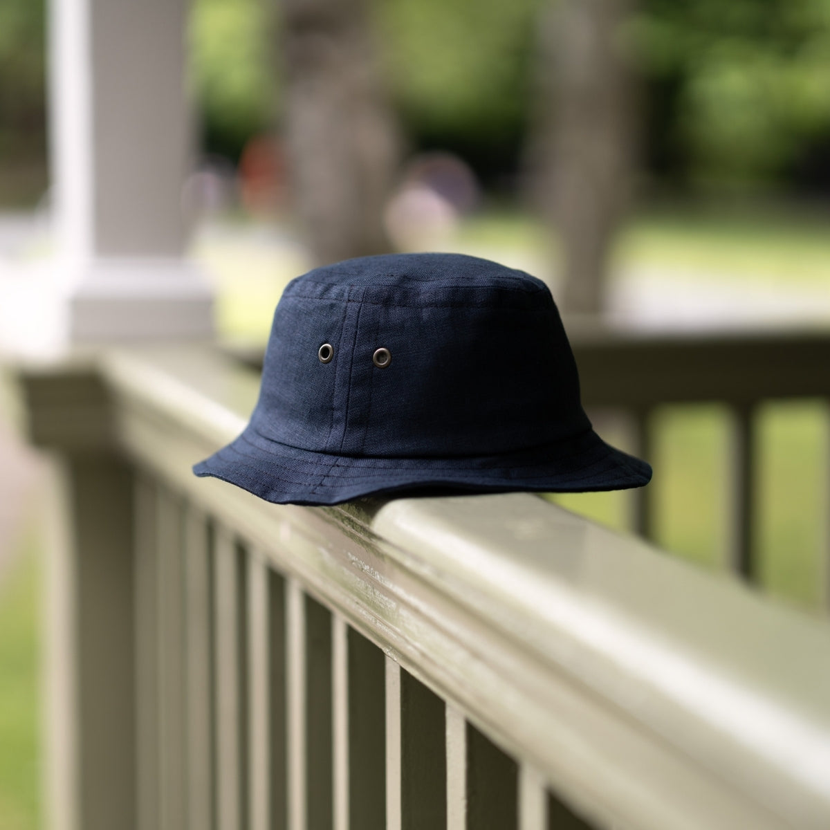 A navy blue bucket hat rests on a light green wooden railing, with a blurred background of trees and greenery in an outdoor setting.