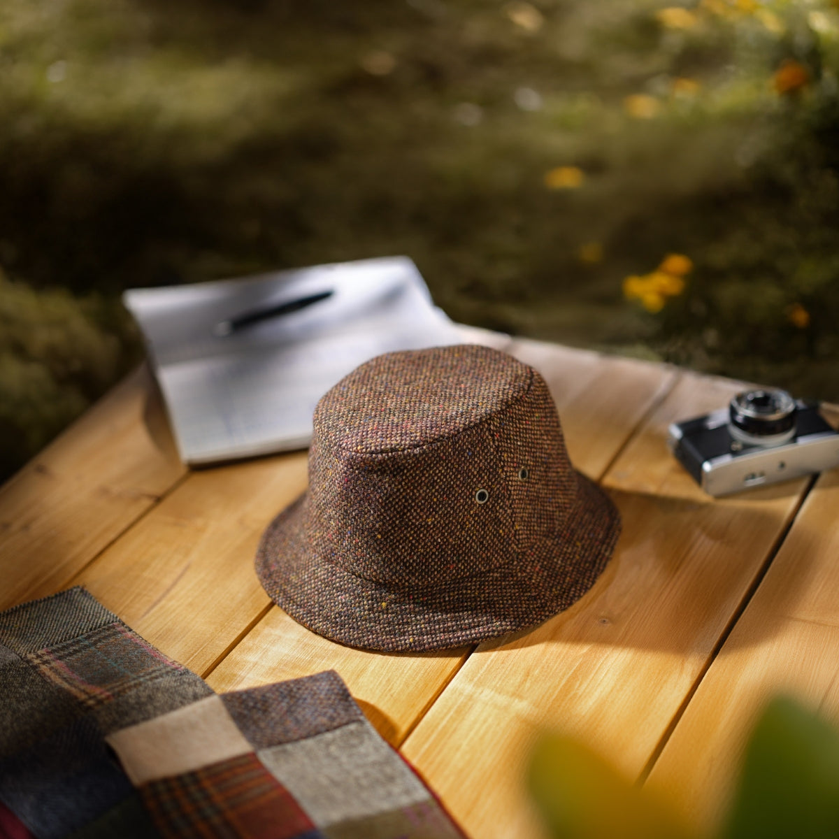 A brown tweed bucket hat sits on a wooden surface next to a patchwork scarf, a pen and notebook, and a vintage camera, with greenery and yellow flowers blurred in the background.