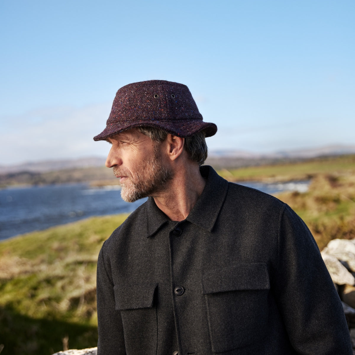 A man with a gray beard wearing a dark jacket and a maroon hat stands outdoors by a body of water, looking to the side, with grassy fields and a blue sky in the background.
