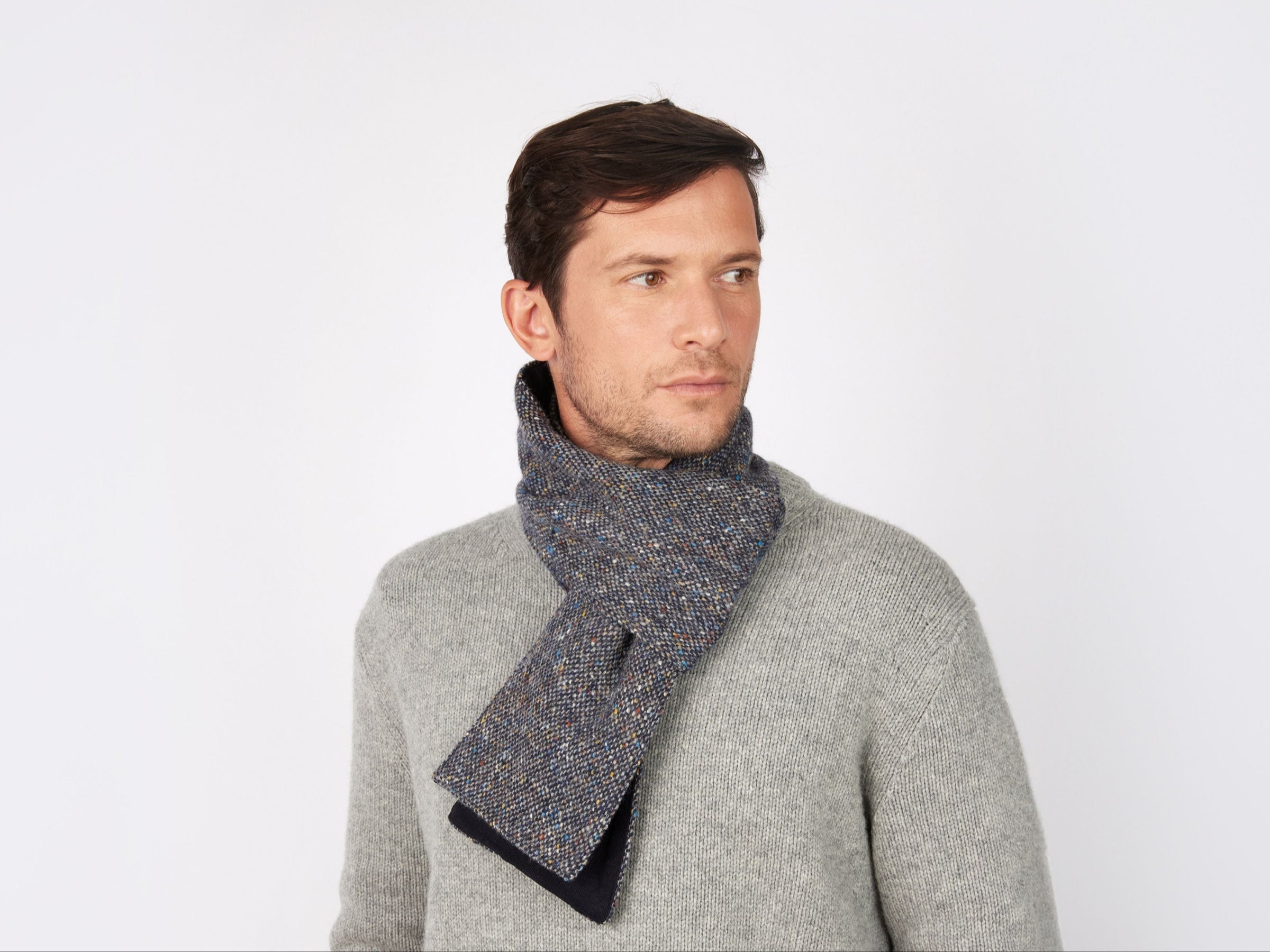 A man in a grey sweater and a Hanna Hats Scarf Tweed / Corduroy, handcrafted in Ireland, stands against a white background, looking slightly left with a neutral expression.