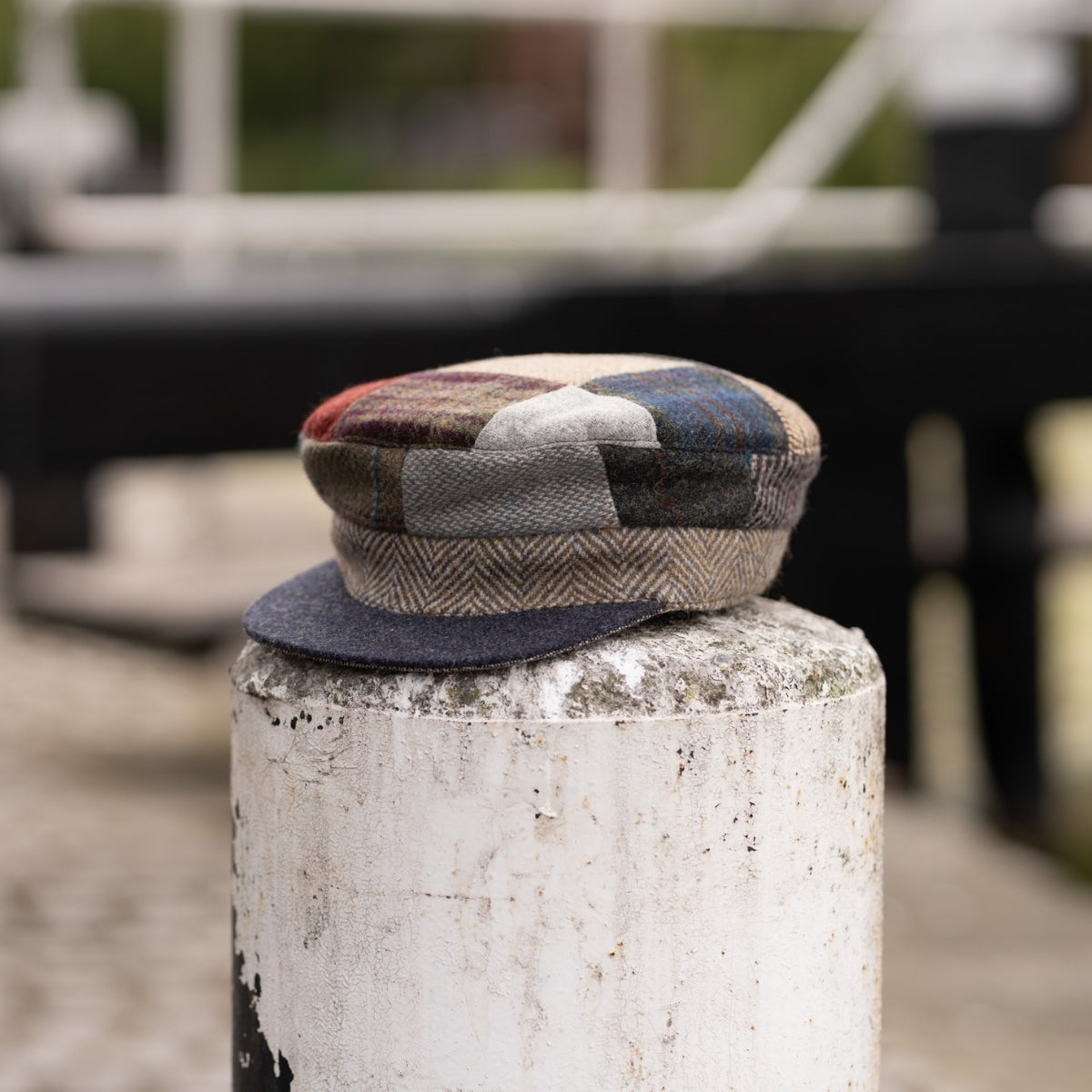 A patchwork flat cap with various fabric patterns and colors sits on top of a weathered white post, with a blurred outdoor background.
