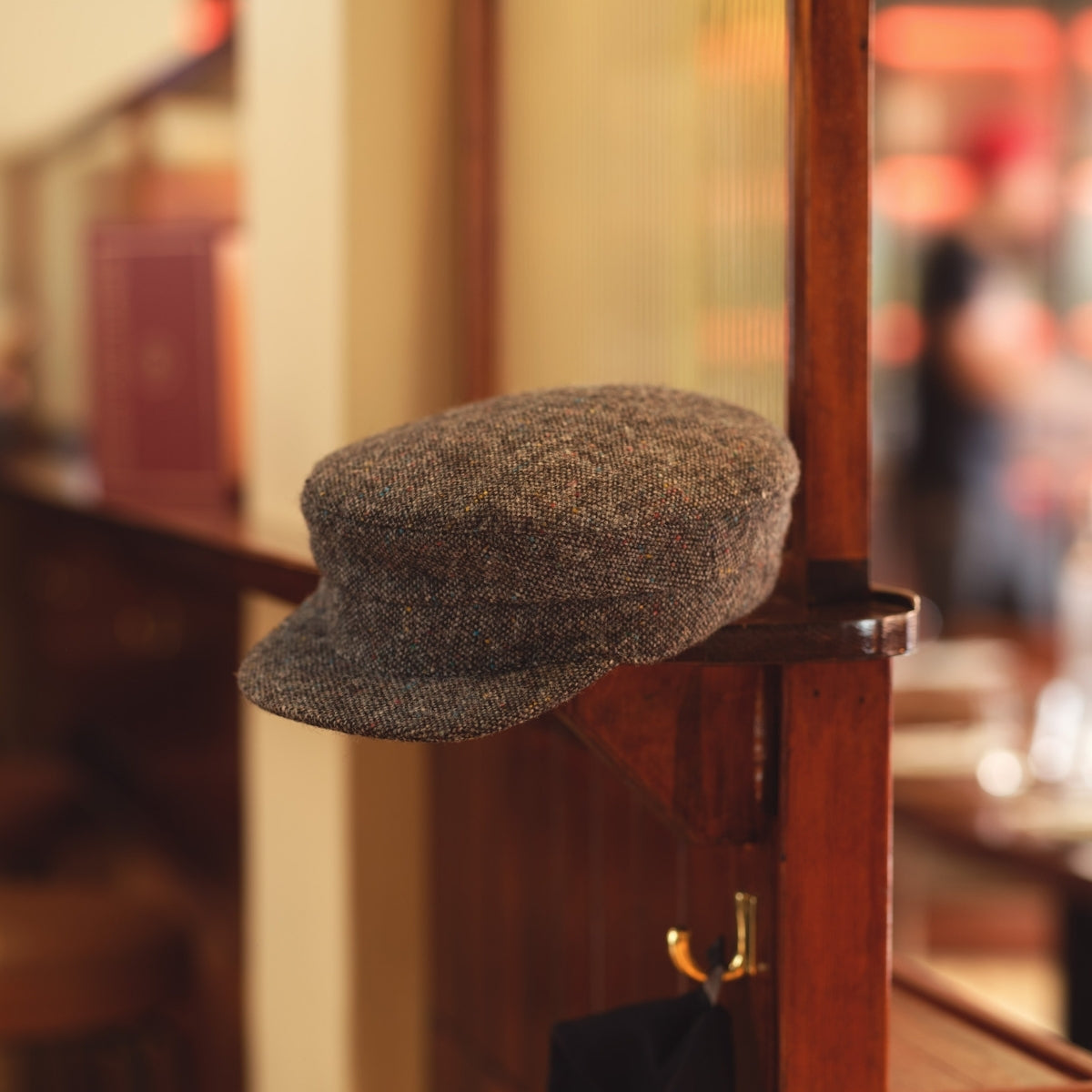 A brown tweed flat cap rests on the corner of a wooden partition inside a warmly lit café or restaurant, with blurred tables and chairs in the background.