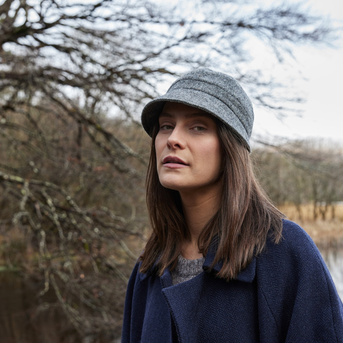 A woman with straight brown hair wearing a grey hat and navy coat stands outdoors near bare trees and a pond, looking calmly at the camera.