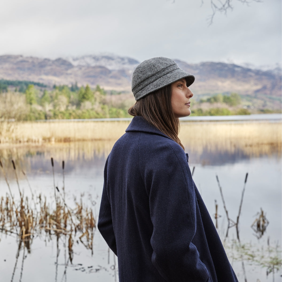 A woman in a gray hat and navy coat stands by a lake, surrounded by reeds, with distant mountains and a cloudy sky in the background.