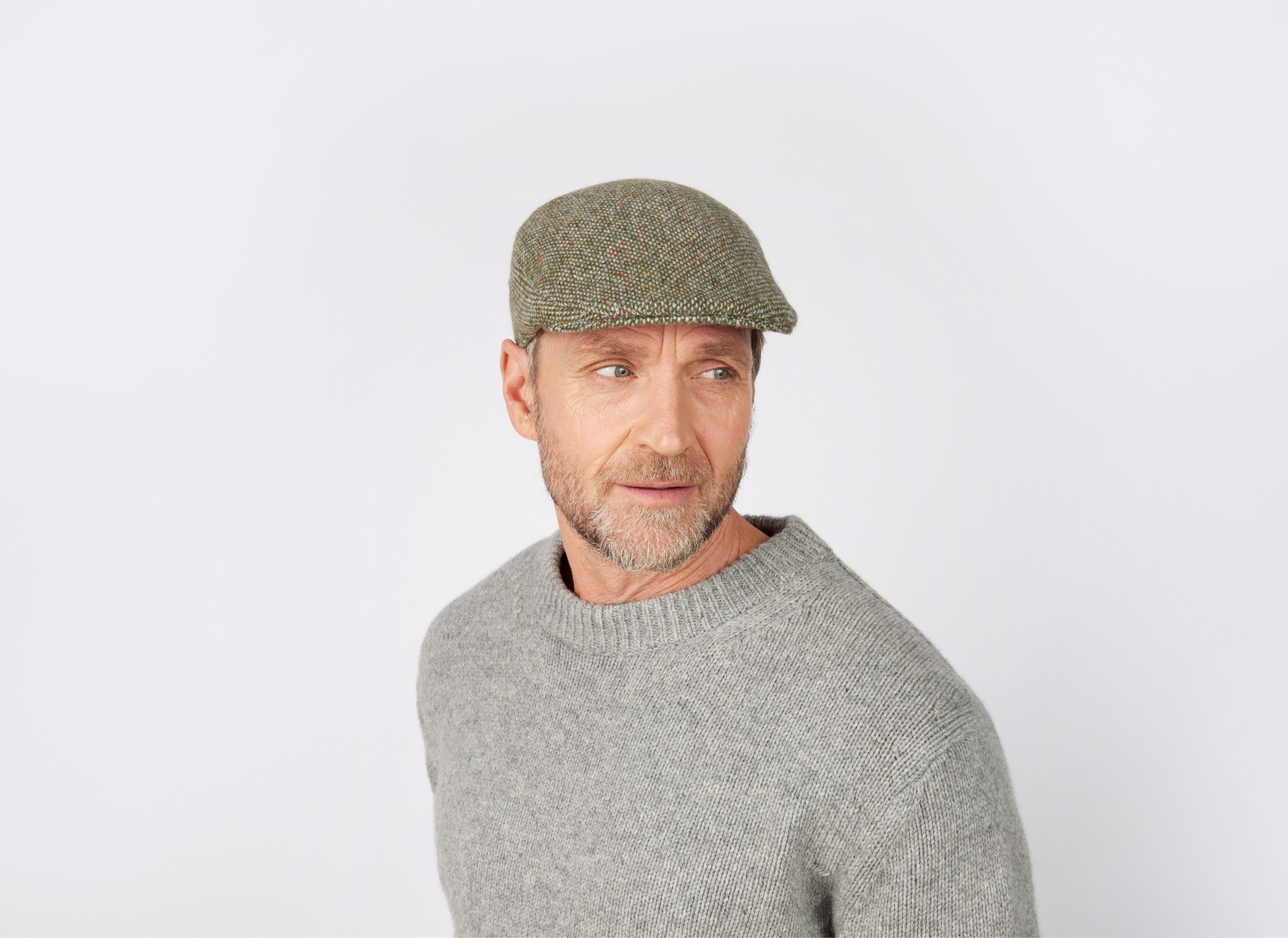 A middle-aged man with a beard, in a gray sweater and the HannaHats Tailor Cap Tweed in green, looks slightly to the side against a plain white background.
