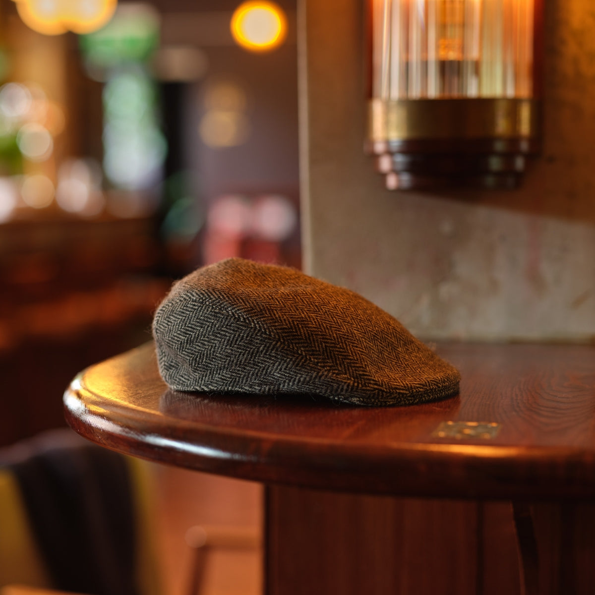 A herringbone flat cap rests on a polished wooden table in a warmly lit, cozy café or pub with blurred lights and furniture in the background.