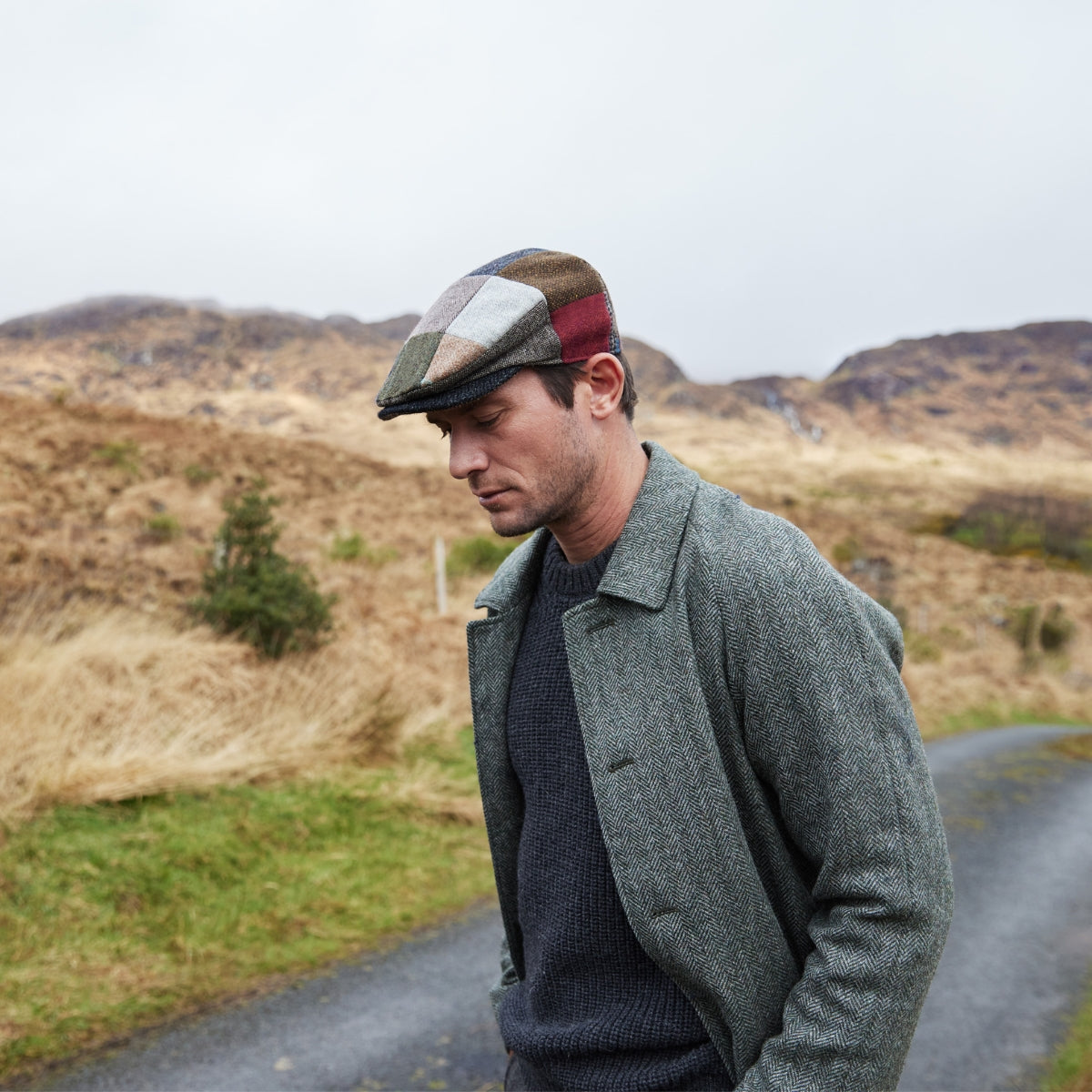 A man wearing a grey jacket, dark sweater, and a colorful patchwork flat cap walks outdoors on a rural road with grassy hills and overcast skies in the background.