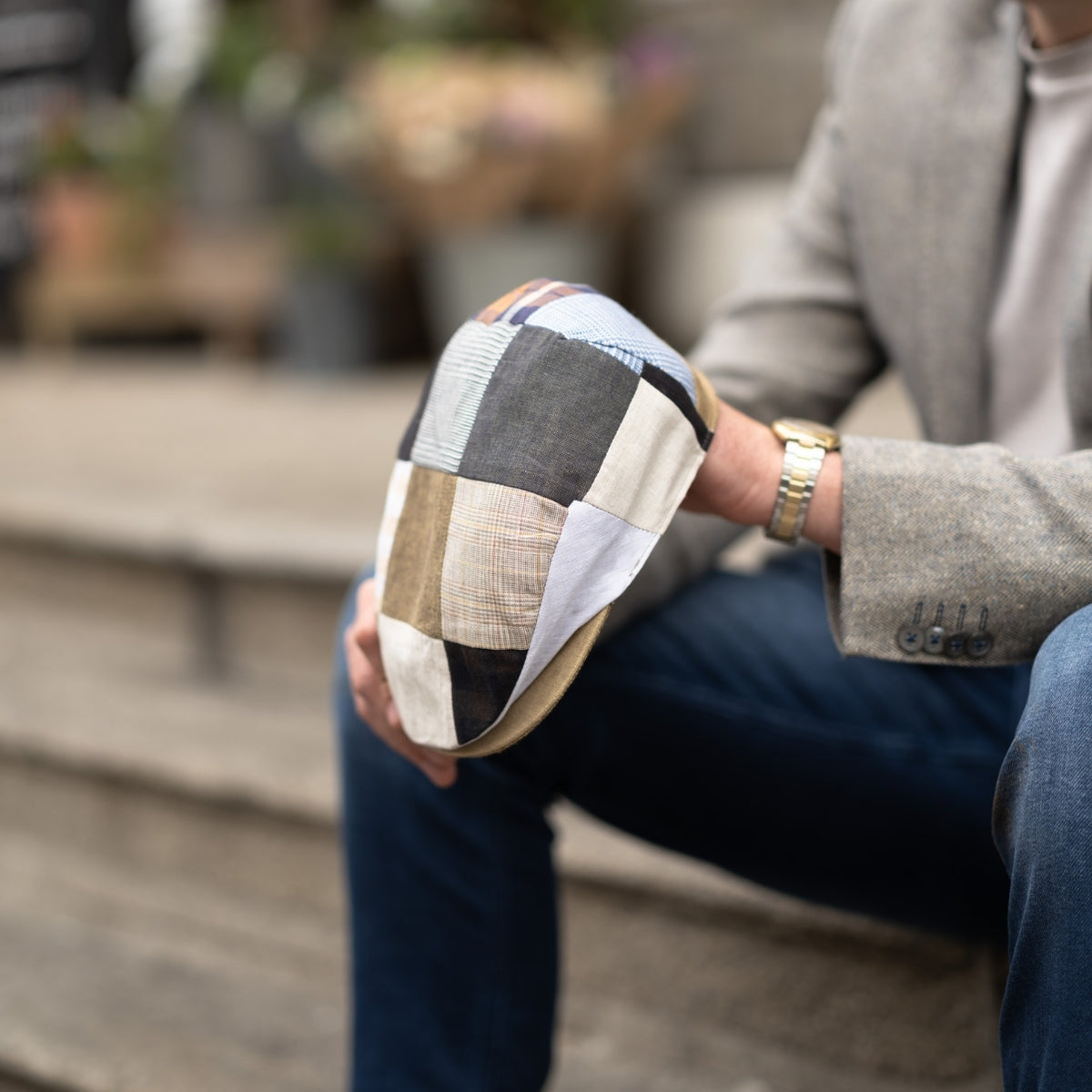 A person in a gray blazer and jeans sits on outdoor steps, holding a patchwork flat cap made of various fabric patterns and colors in their hand. The focus is on the cap; the person's face is not visible.