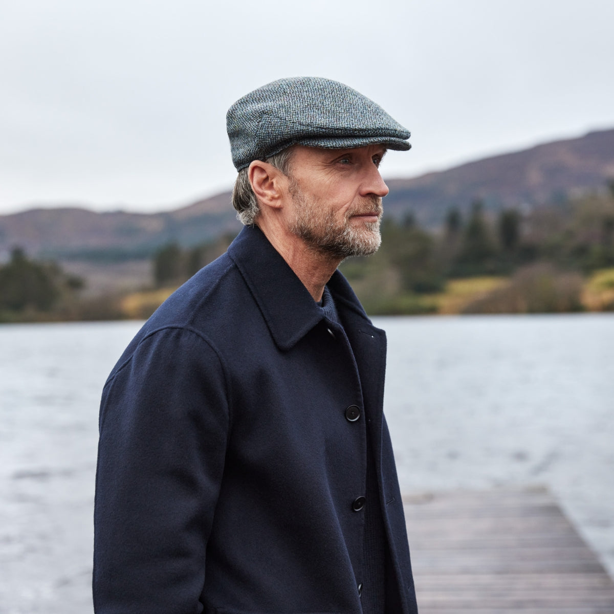A man with a gray beard wearing a flat cap and dark coat stands near a lake, with hills and trees in the background on a cloudy day.